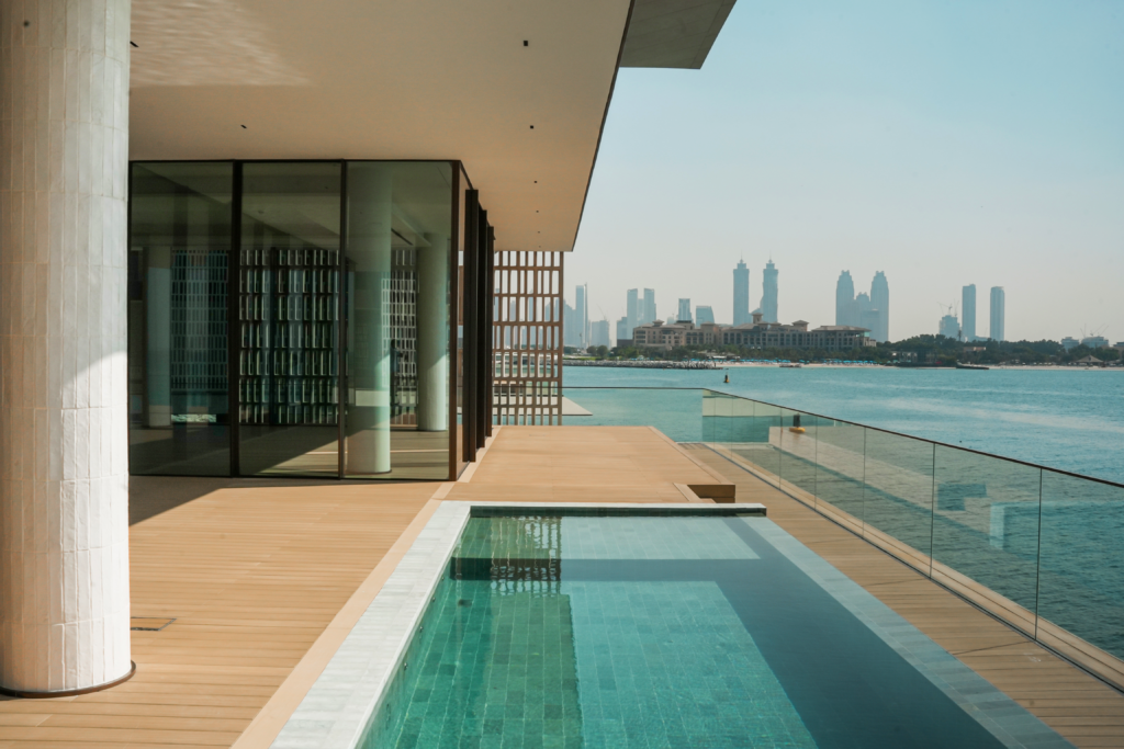 Pale deck alongside a lap pool with a frameless glass balustrade, a vertical timber slat screen at one end and a city skyline across the water