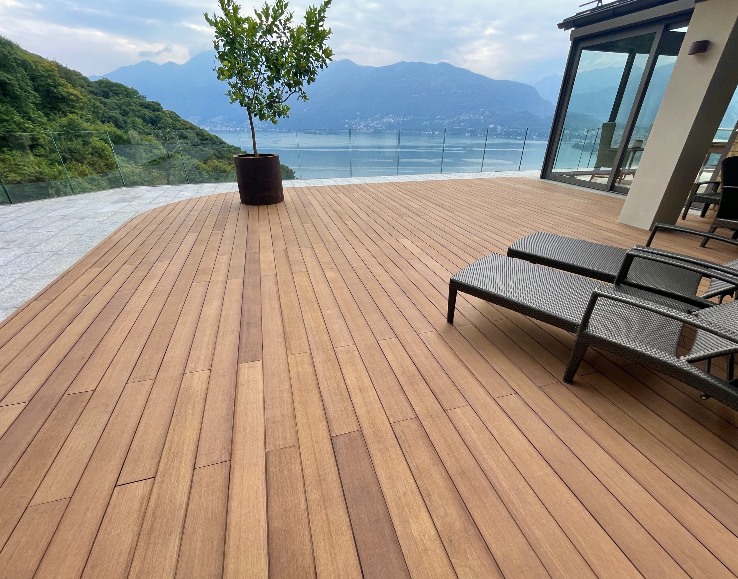 Warm bamboo terrace with sun loungers and a potted tree, glass balustrade, lake and mountains beyond