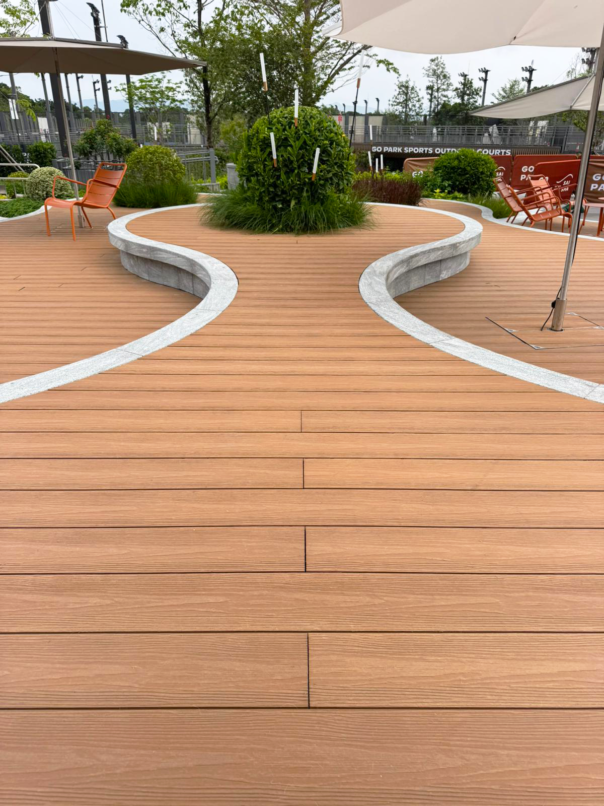 Warm composite deck with curved stone bands set into it and built-in curved benches around a planted island, parasols overhead