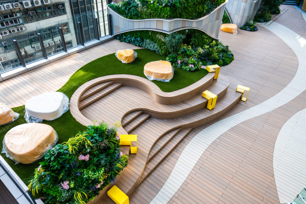 Rooftop garden terrace with curved decking in two tones, a curved timber bench, planted beds and yellow stools, city buildings behind