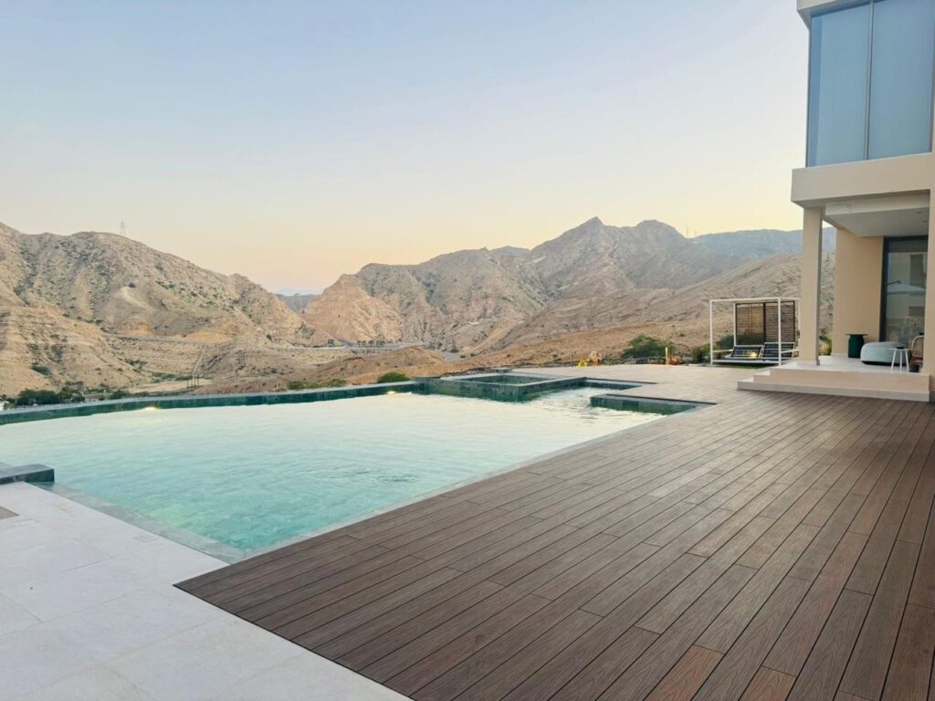 Infinity pool framed by dark brown composite decking at a hillside villa, mountains at dusk beyond the water