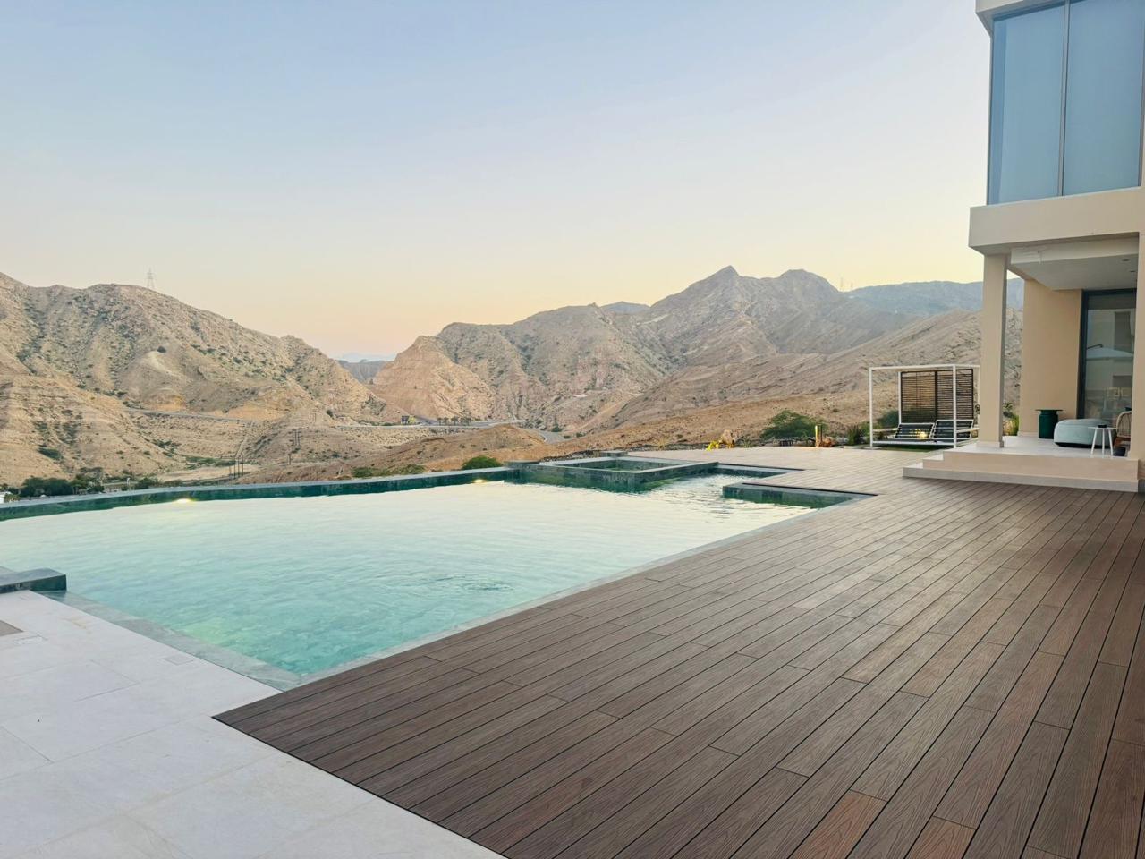 Infinity pool framed by dark brown composite decking at a hillside villa, mountains at dusk beyond the water