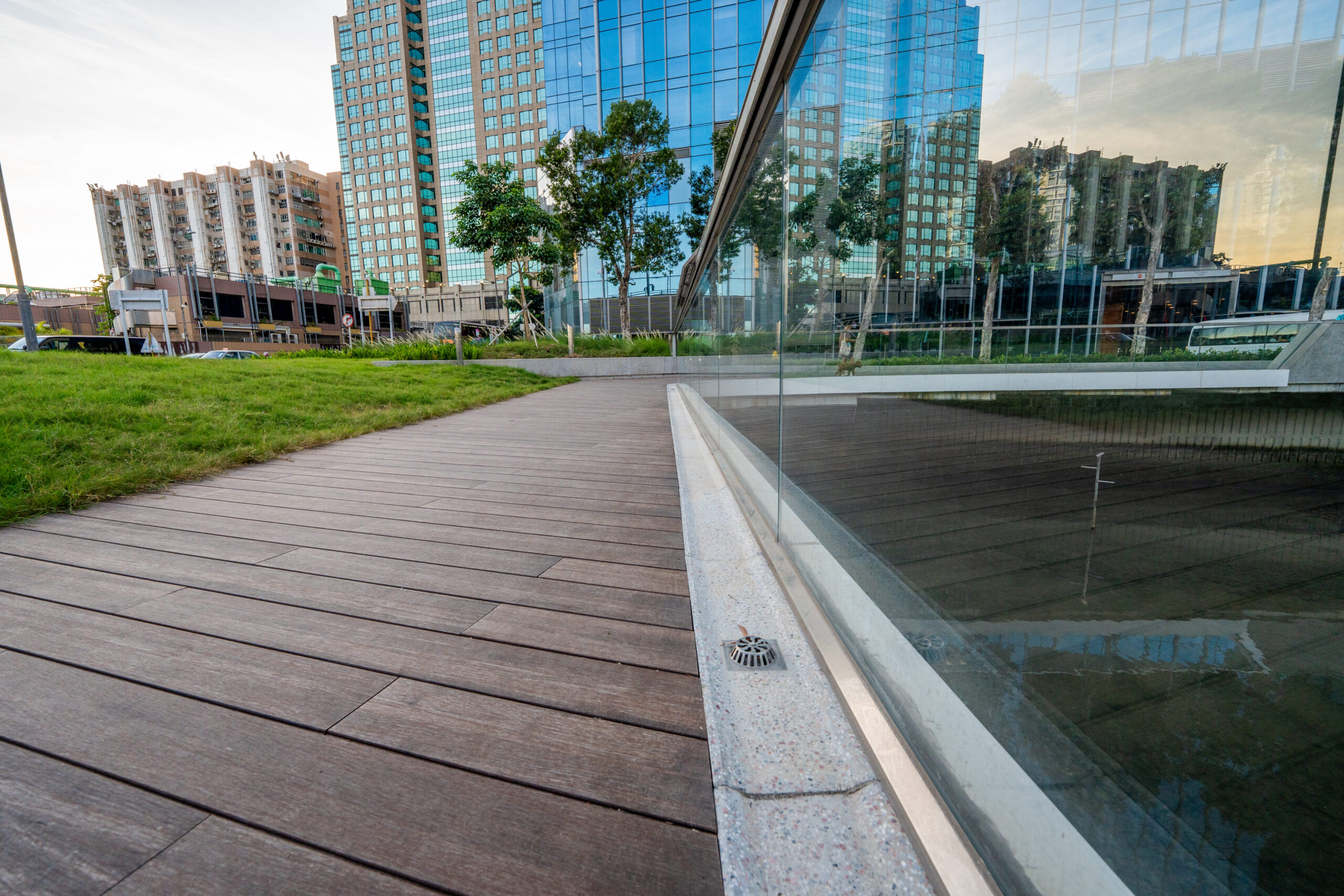 Dark composite decking meeting a frameless glass balustrade at the edge of a reflecting pool, city towers behind