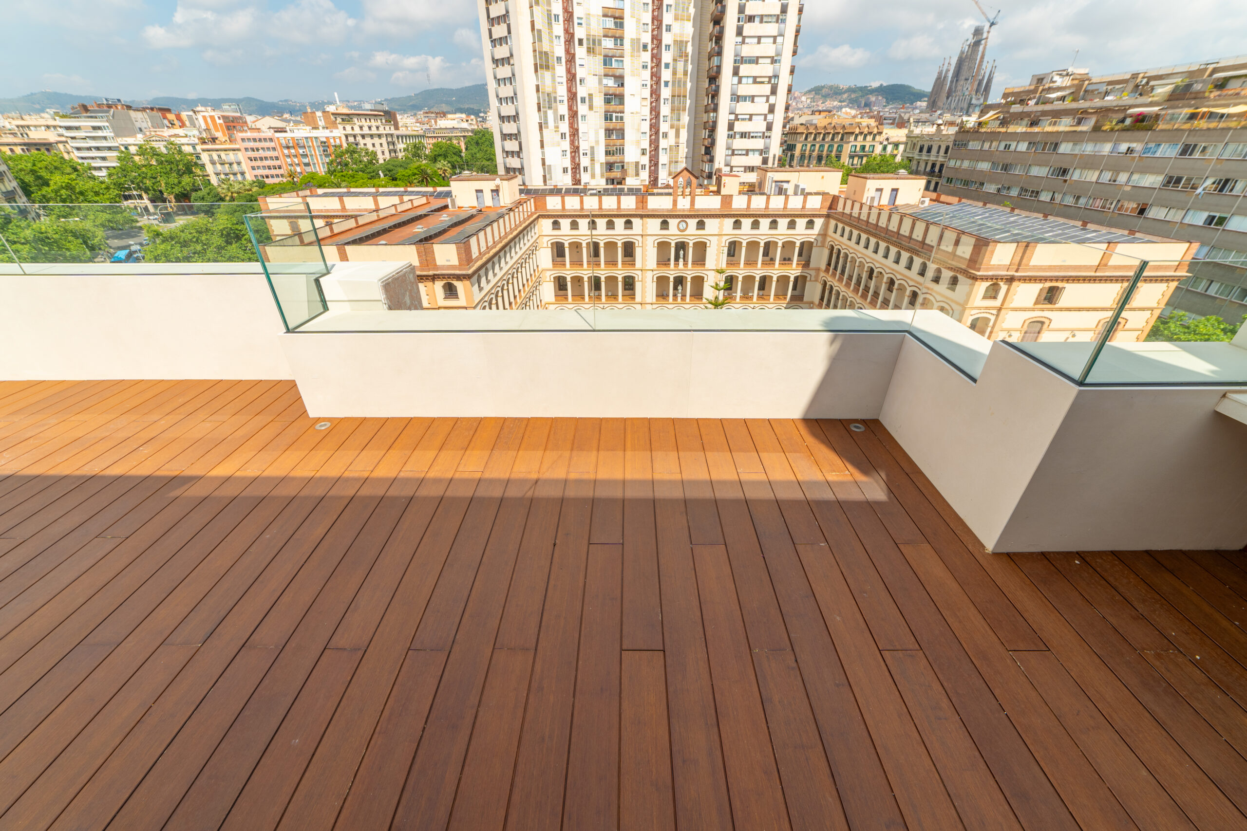 Compact rooftop terrace in warm hardwood decking with a low white parapet topped by glass, city rooftops beyond