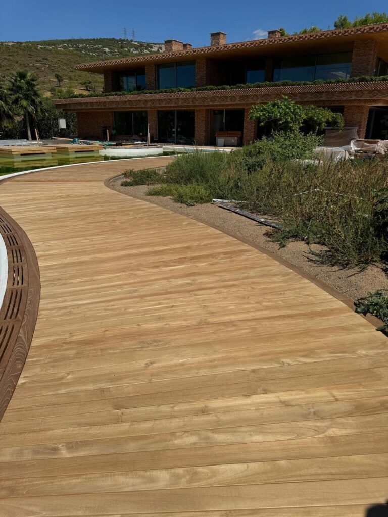 Curved pale hardwood deck path running past planting toward a low modern brick house on a hillside
