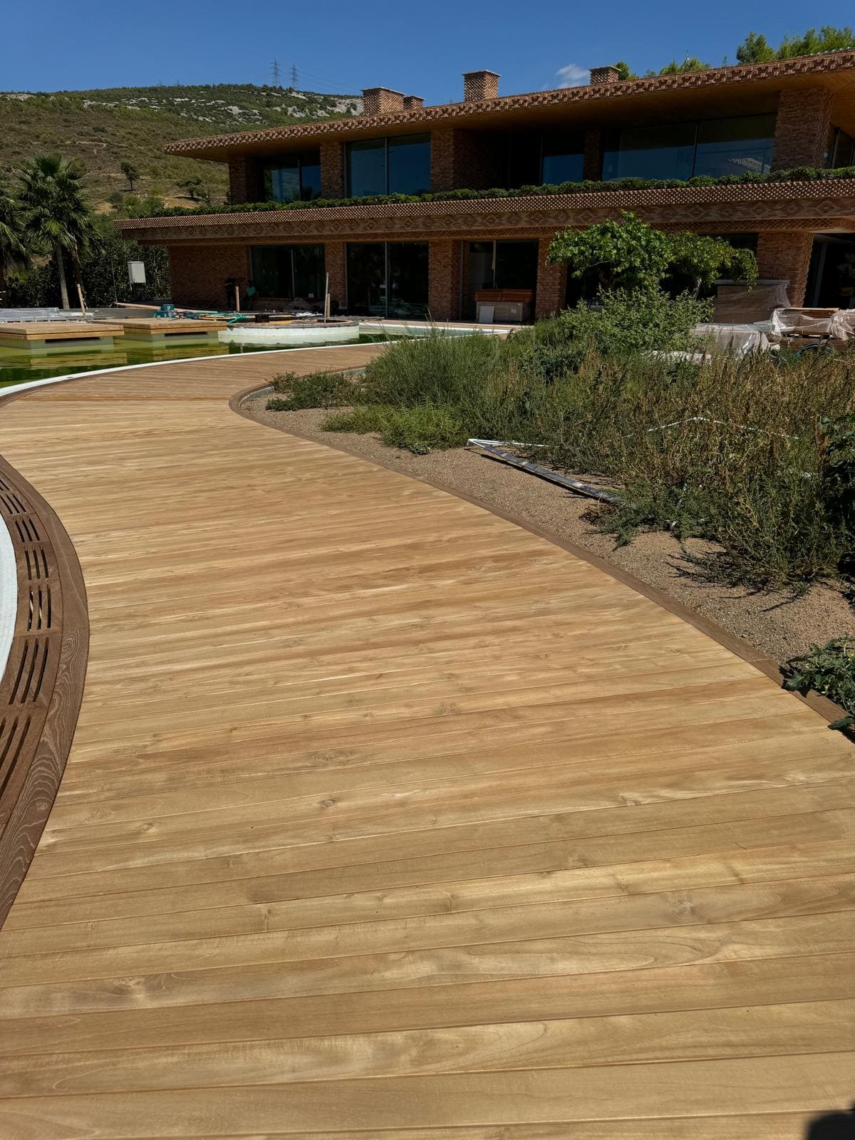 Curved pale hardwood deck path running past planting toward a low modern brick house on a hillside