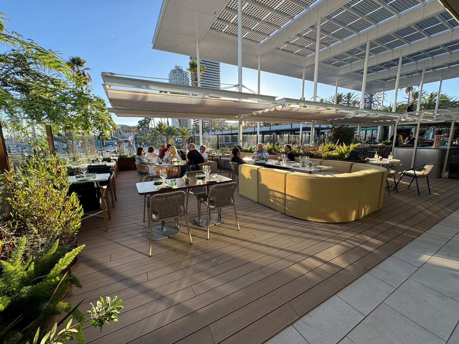 Restaurant terrace under a slatted pergola, with dining tables on composite decking meeting pale stone paving, planting along the edge